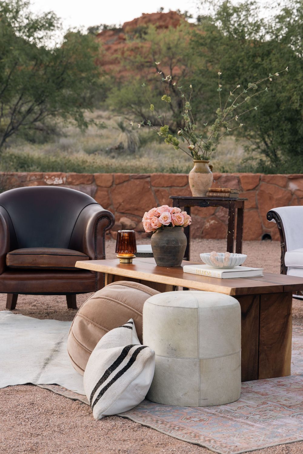 Cozy outdoor patio setup with brown leather chair, cream poufs, and pink flowers in vase against desert landscape backdrop