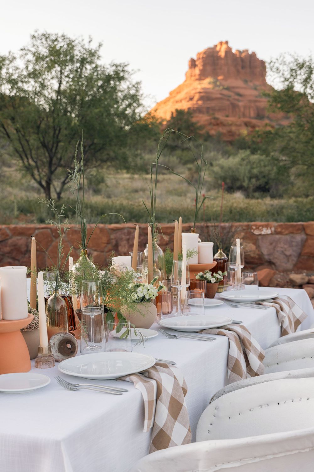 An elegant outdoor table setting with white linens and candles against a red rock formation in Sedona at sunset.