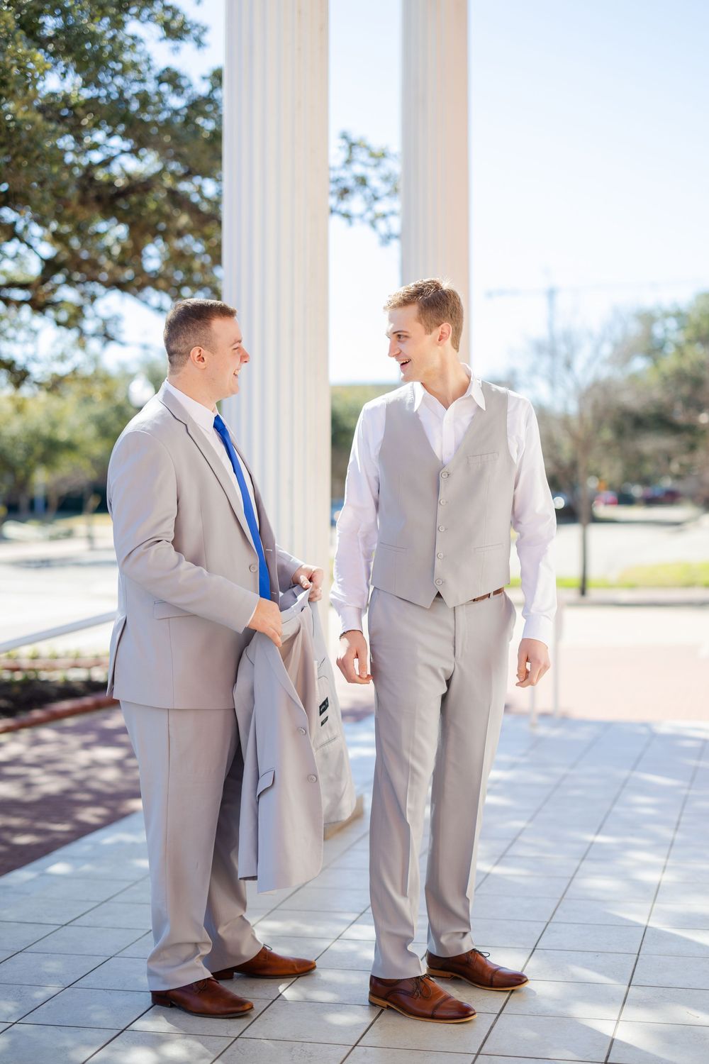 Two people in light gray suits and brown shoes converse on a columned porch.