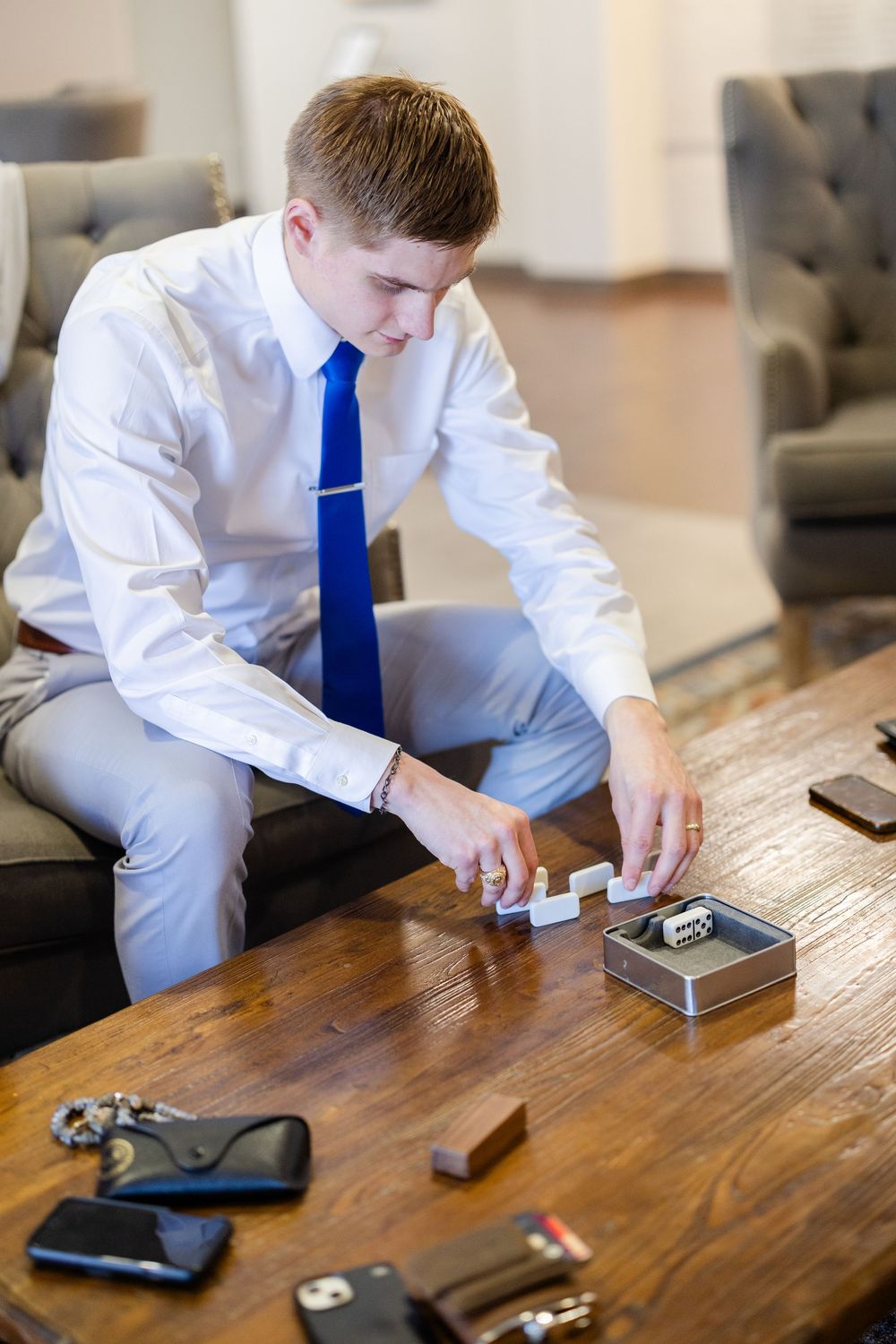 Groom in white shirt and blue tie arranges wedding rings and details on wooden table during wedding preparations.