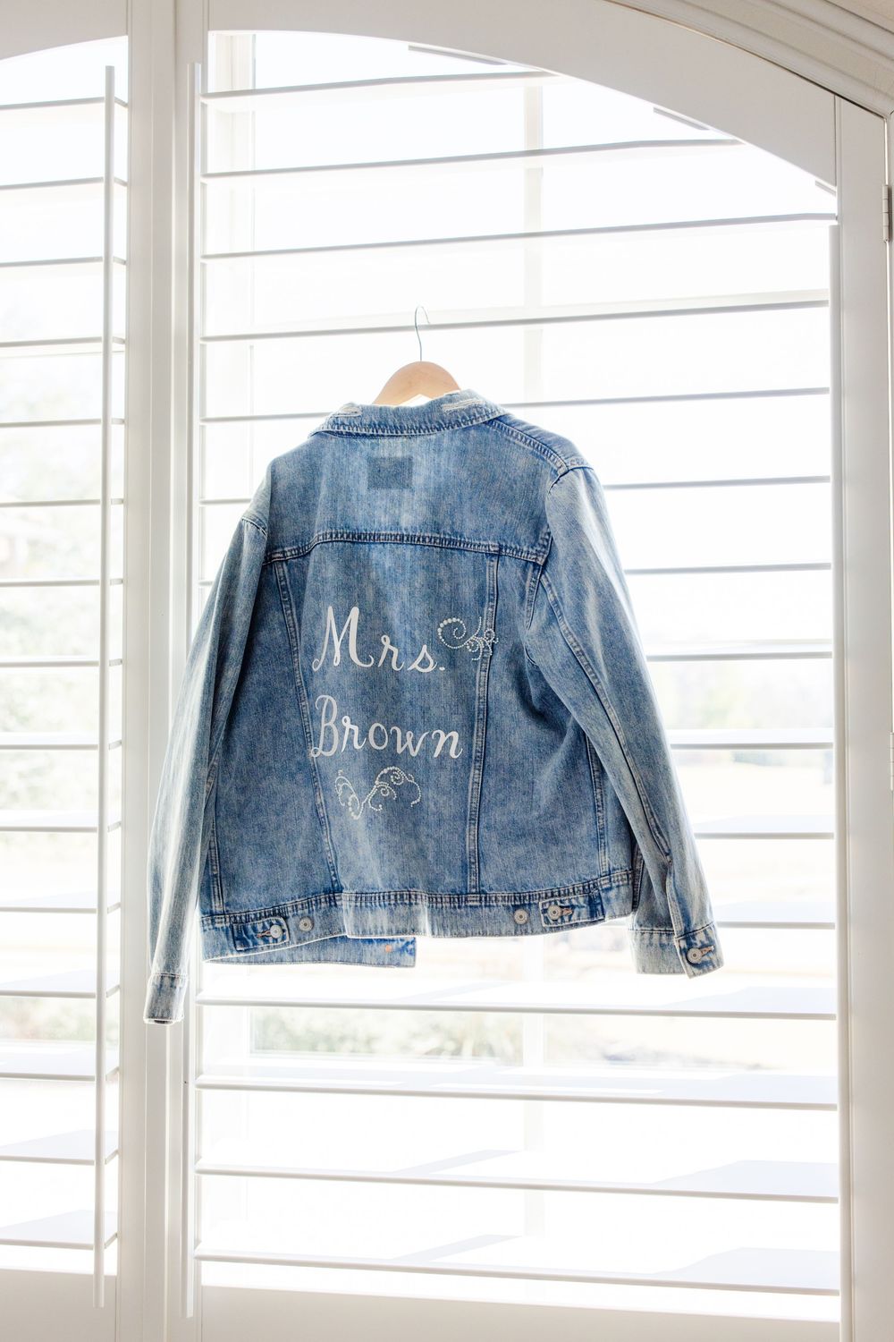 A customized denim jacket with 'Mrs. Brown' embroidered text hangs on display against white shutters.