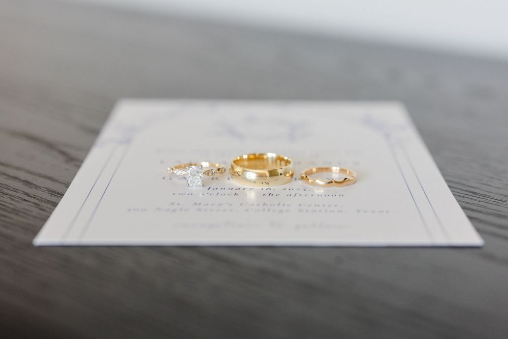 Wedding rings and invitation details displayed on a dark wooden surface.
