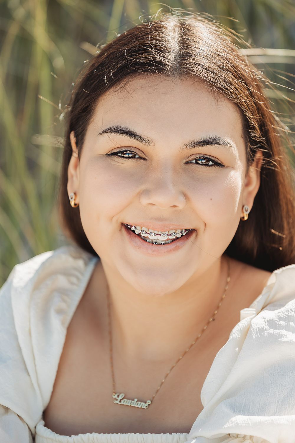Portrait of a smiling young woman in white wearing braces and necklace with foliage background in natural lighting.