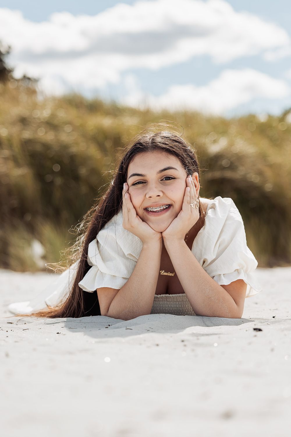Person in white top lying on sand at the beach with hands cupping face on a sunny day with soft cloudy sky in background.