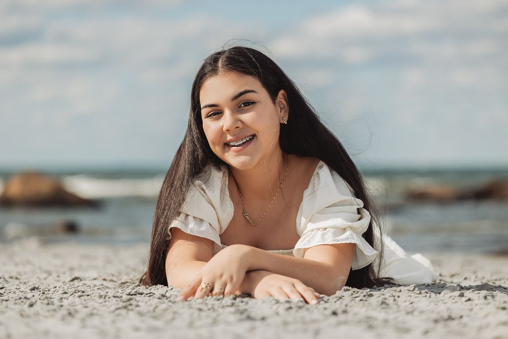 Person lying on sandy beach near ocean with a relaxed expression wearing a white top in natural sunlight.