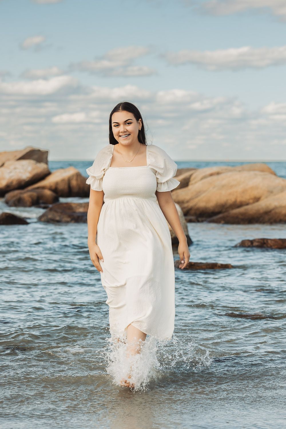 A figure in a flowing white dress splashes through ocean waves near rocky coastal formations at sunset.