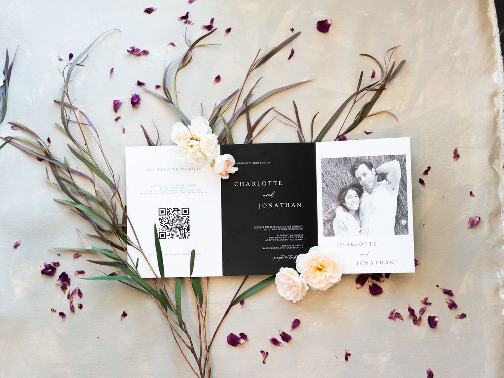 Wedding invitation cards in black and white with QR code arranged with delicate flowers and rose petals on neutral fabric.