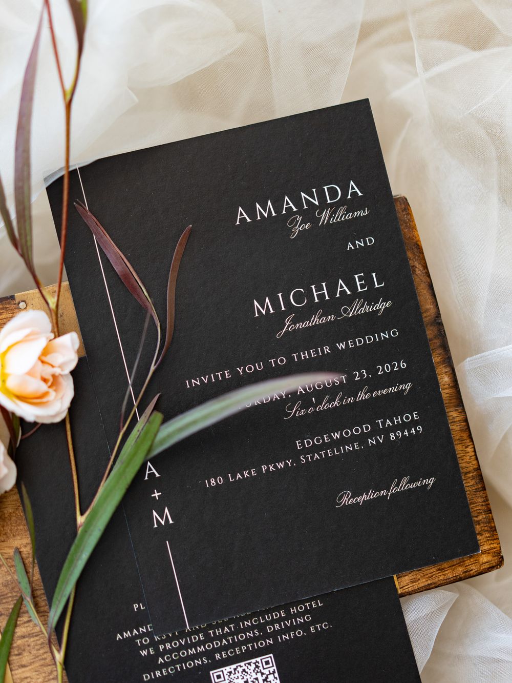 Elegant black wedding invitation with white text and minimalist floral design displayed on a wooden surface with roses.
