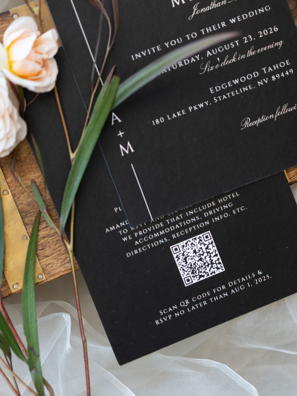 Elegant black invitation cards with QR codes displayed alongside white roses and eucalyptus greenery on marble surface.