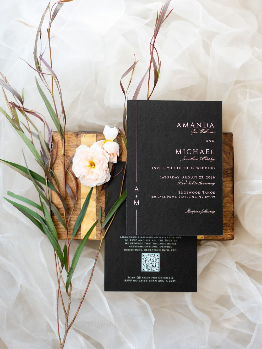 Elegant black wedding invitation with white text displayed alongside white orchids and greenery on a light fabric background.