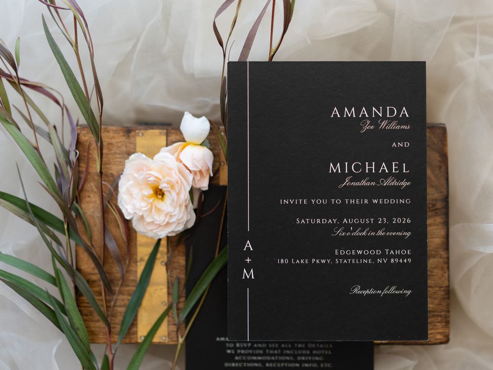 Modern black wedding invitation with white text displayed alongside pink roses and wooden decor elements.