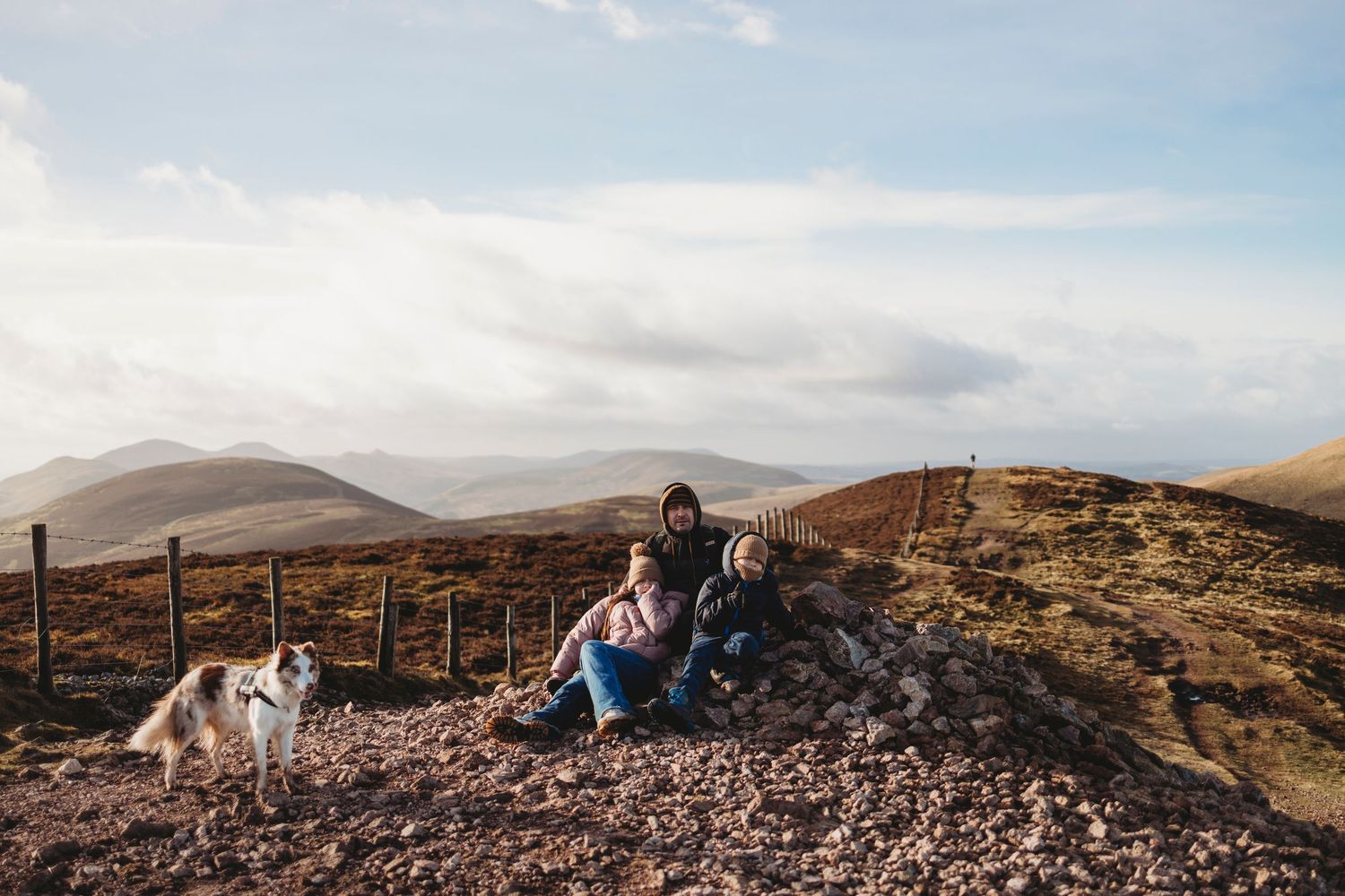Hiker rests on rocky mountain trail with white dog while taking in scenic views of rolling hills and cloudy skies.