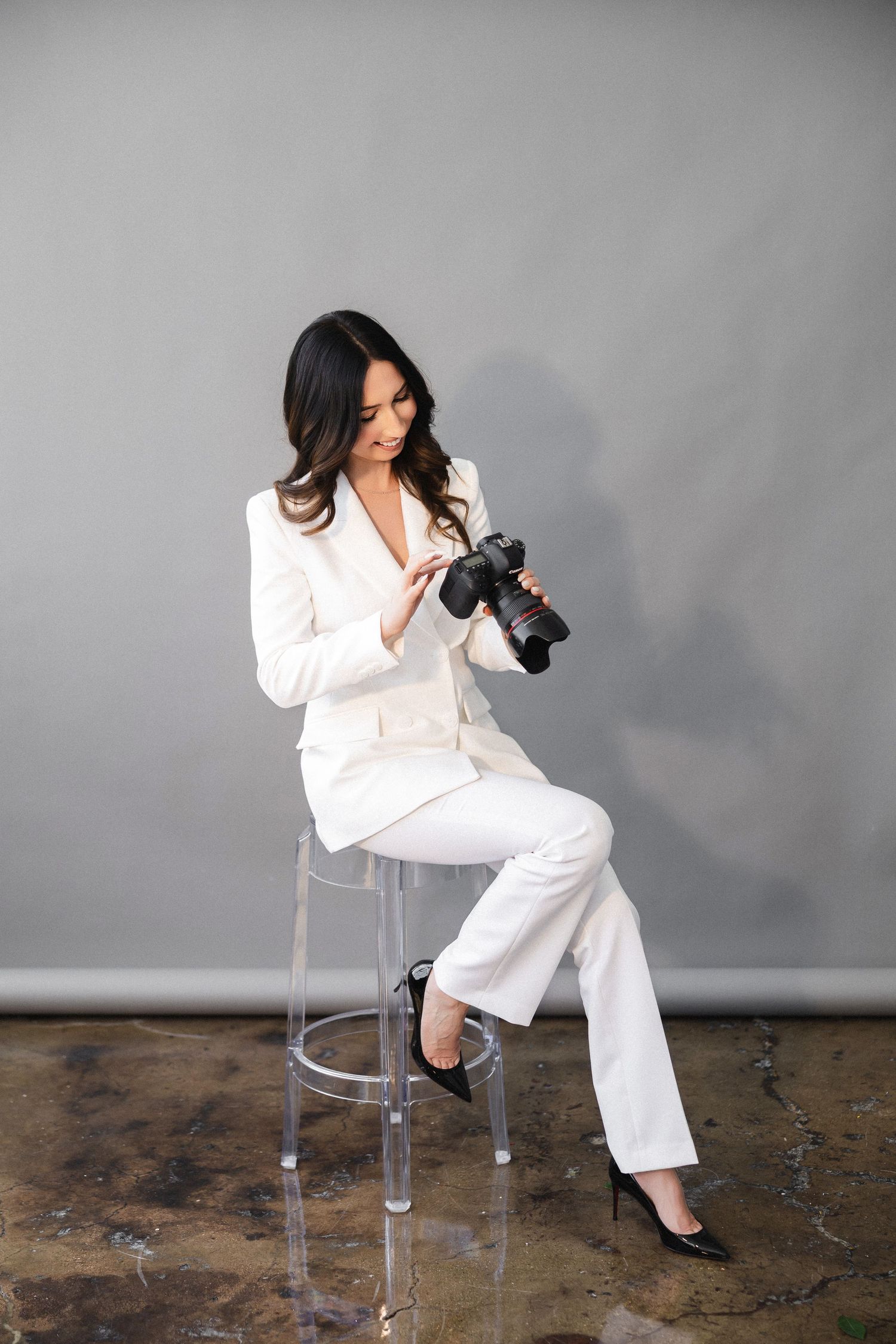 Professional photographer in crisp white suit reviews photos on camera while seated on clear acrylic stool against grey wall.