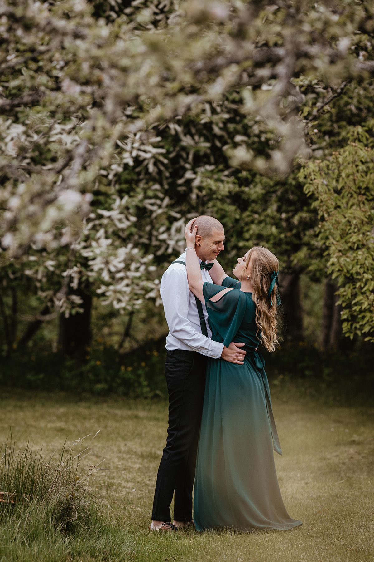 Vackert brudpar i en grönskande trädgård i maj där äppelträden blommar. De står under trädkronorna och håller om varandra med blommorna ovan