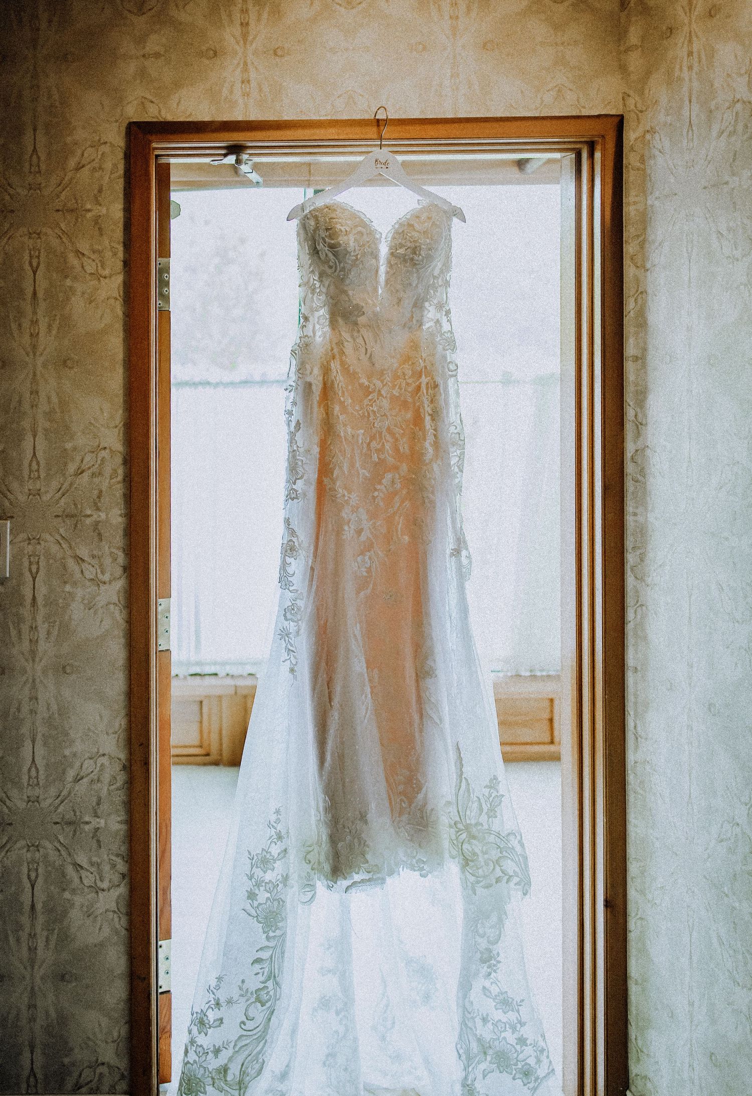 An elegant white lace wedding dress with sheer details hangs displayed in a wooden doorframe against textured walls.