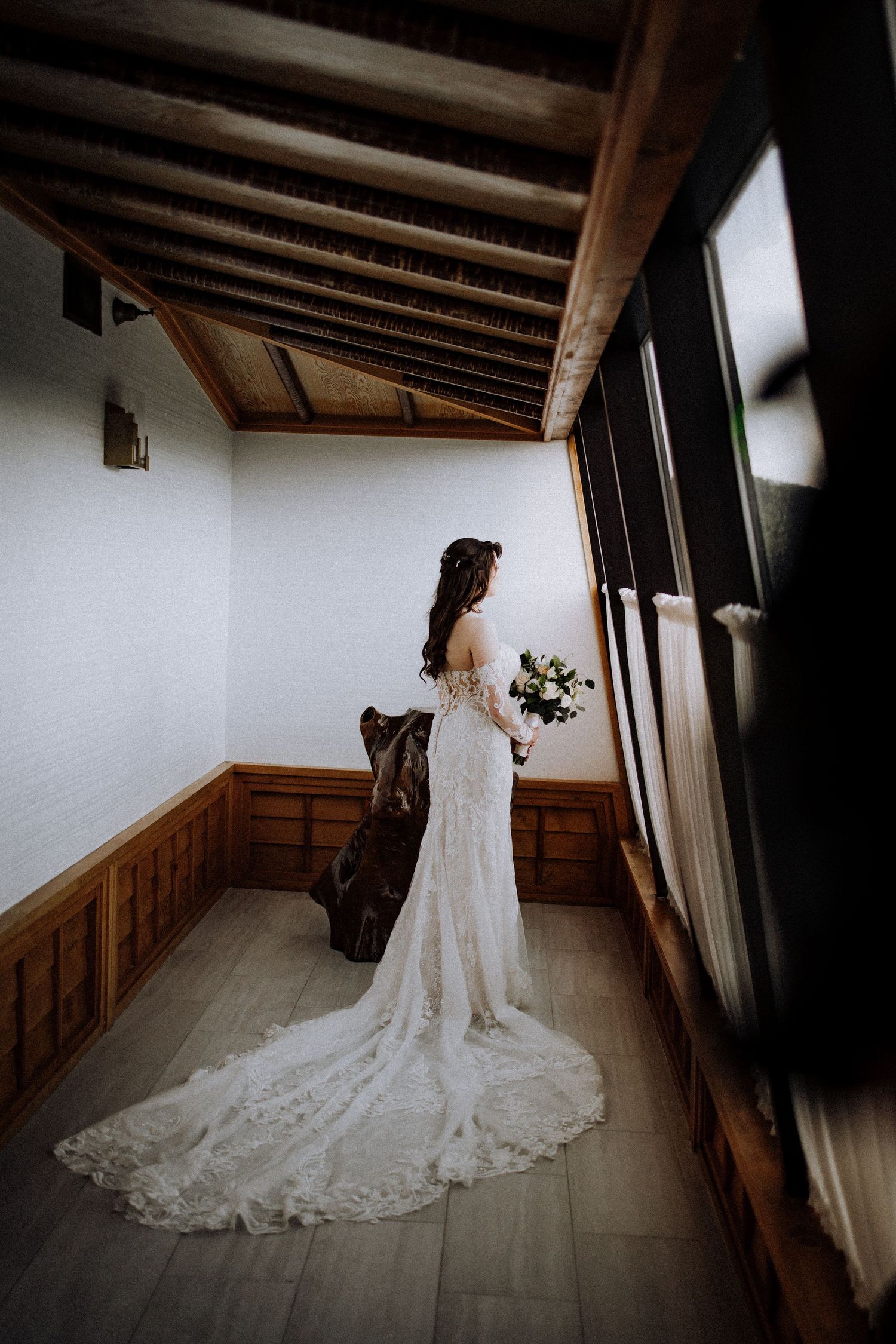 Bride in lace wedding dress with dramatic train stands by window in rustic wood-paneled room holding white bouquet.