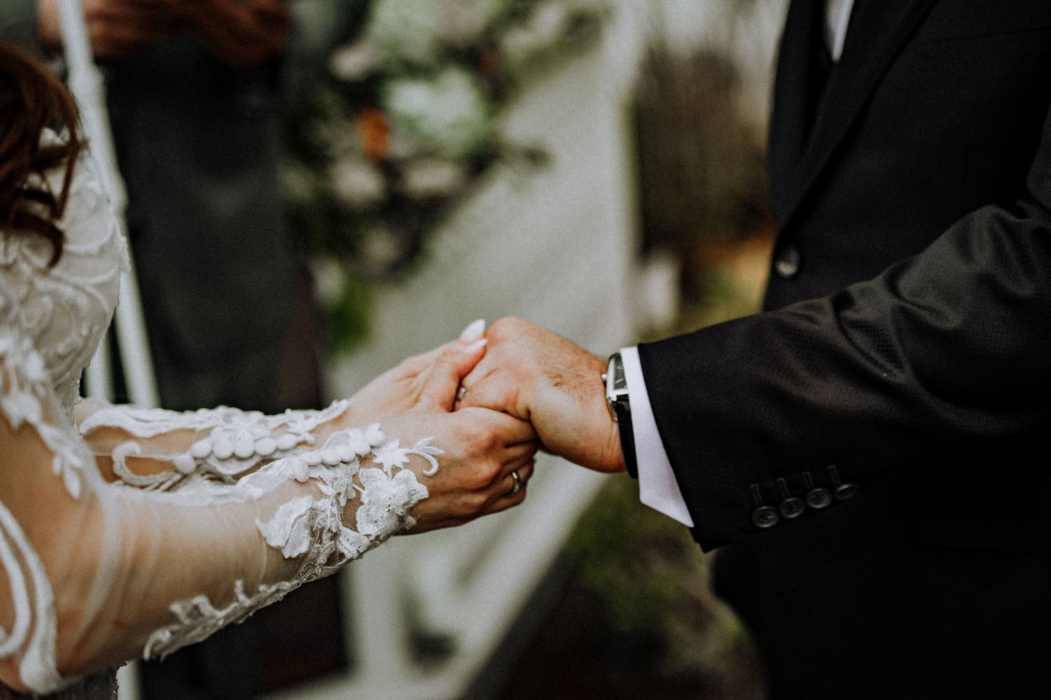 Close-up of bride and groom holding hands during wedding ceremony, showing lace sleeve details and formal attire.