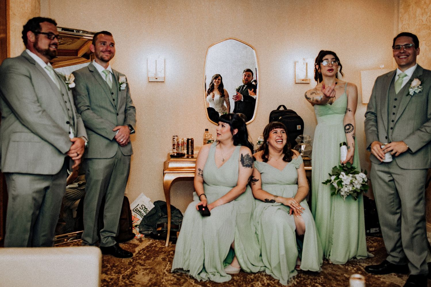 Wedding party in sage green attire gathered during indoor ceremony with soft lighting and floral decor.