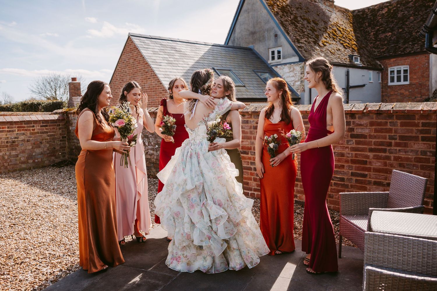 A festive wedding scene with bridesmaids in rust and burgundy dresses gathered joyfully celebrating outside a brick building.