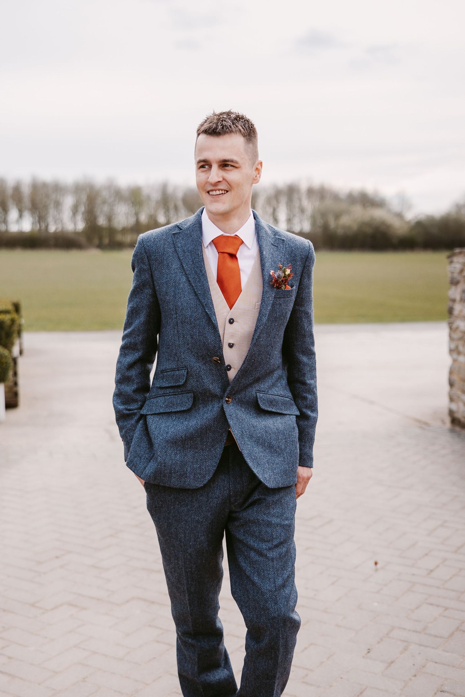 A groom in a stylish blue tweed suit with orange tie and cream waistcoat stands outdoors at a wedding venue.