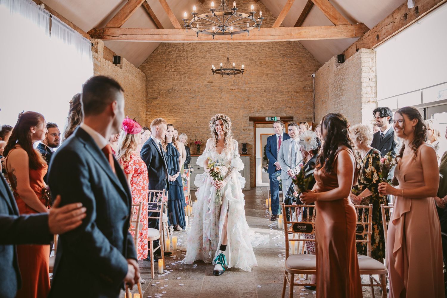 A rustic barn wedding ceremony takes place in a stone-walled venue with wooden beams and a chandelier overhead.