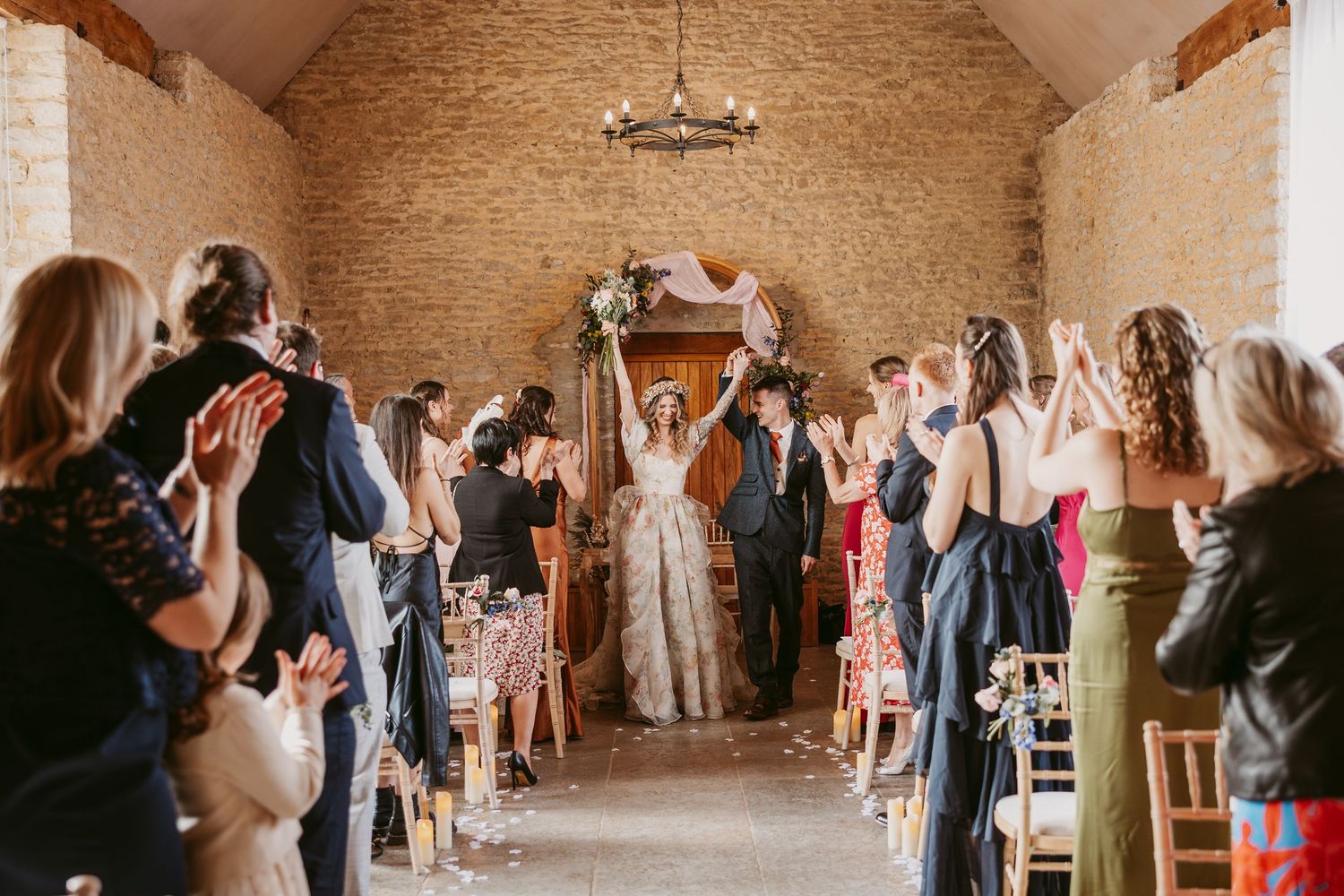 Guests celebrate as newlyweds walk down the aisle inside rustic stone barn during wedding ceremony.