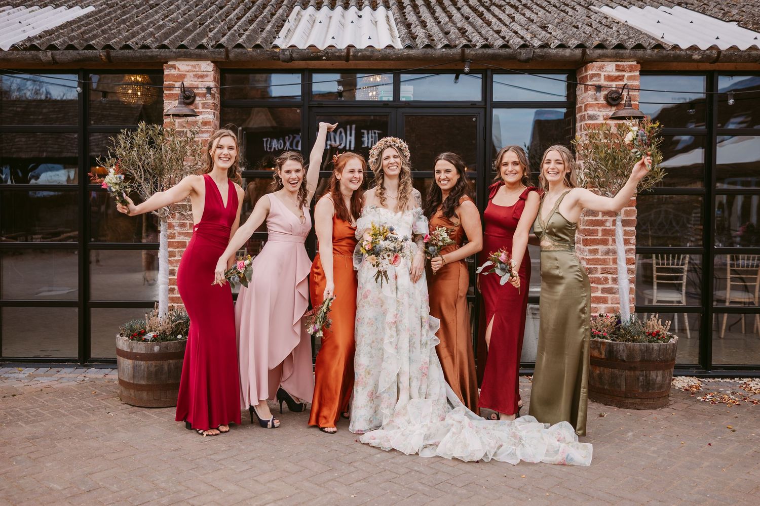 Wedding party in mix-matched formal dresses poses together outside rustic venue with brick columns.