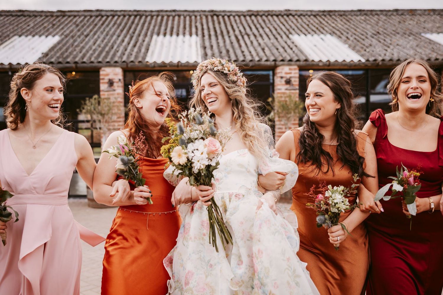 A joyful wedding party wearing colorful pink, rust and burgundy dresses share a candid laughing moment together outdoors.