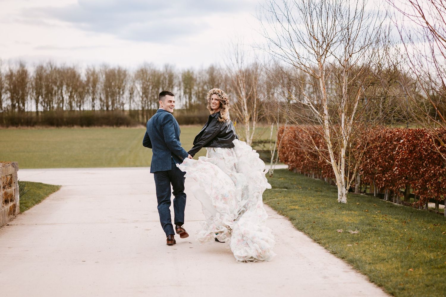 A couple walking down a tree-lined path in a garden setting, with the bride's white dress flowing in the wind.