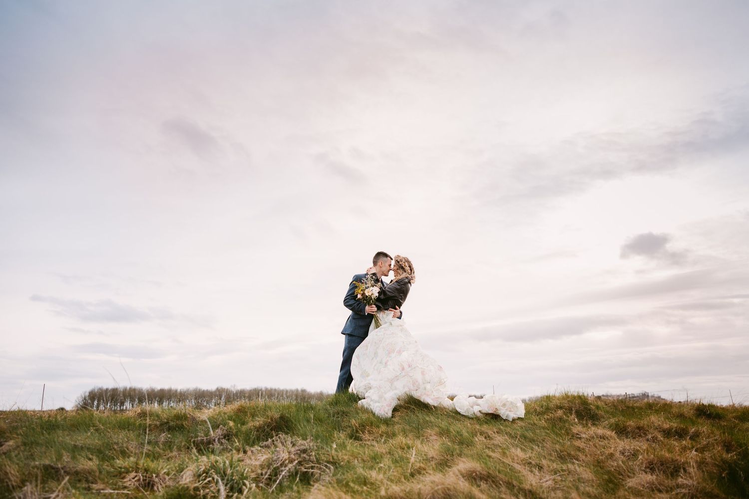 Couple embraces on grassy hilltop against cloudy sky during romantic outdoor photo session.
