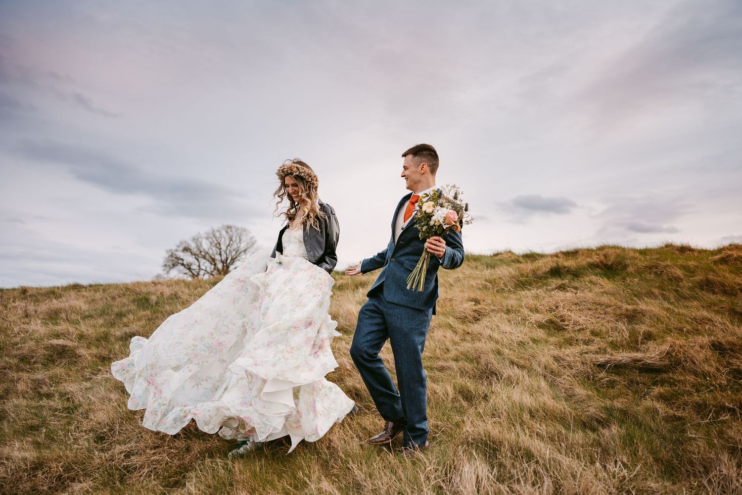 Two people walking across a windswept hilltop field during a wedding photoshoot with dramatic cloudy skies.