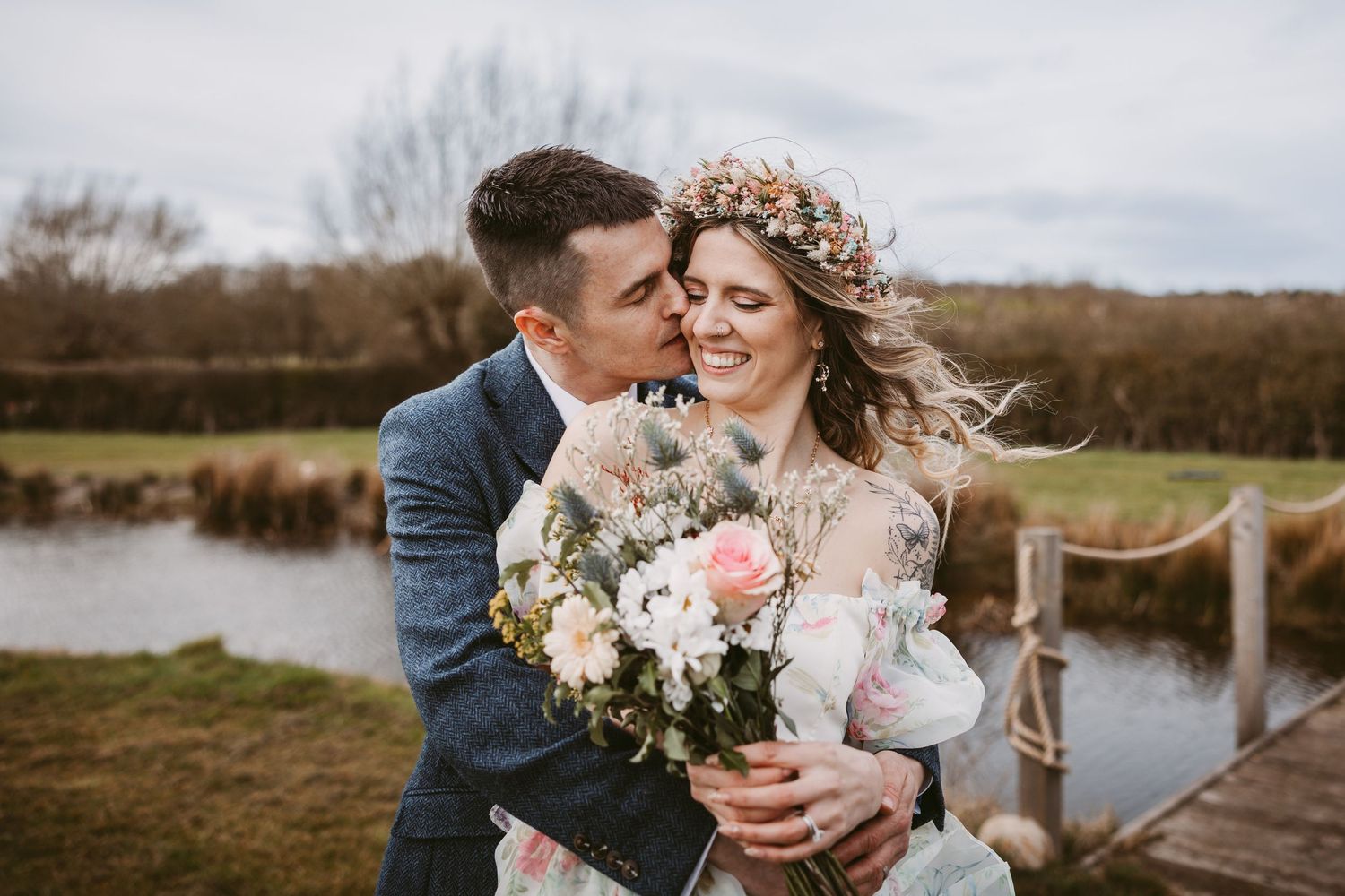 A couple shares an intimate moment at their rustic outdoor wedding by a lake while holding a pastel floral bouquet.
