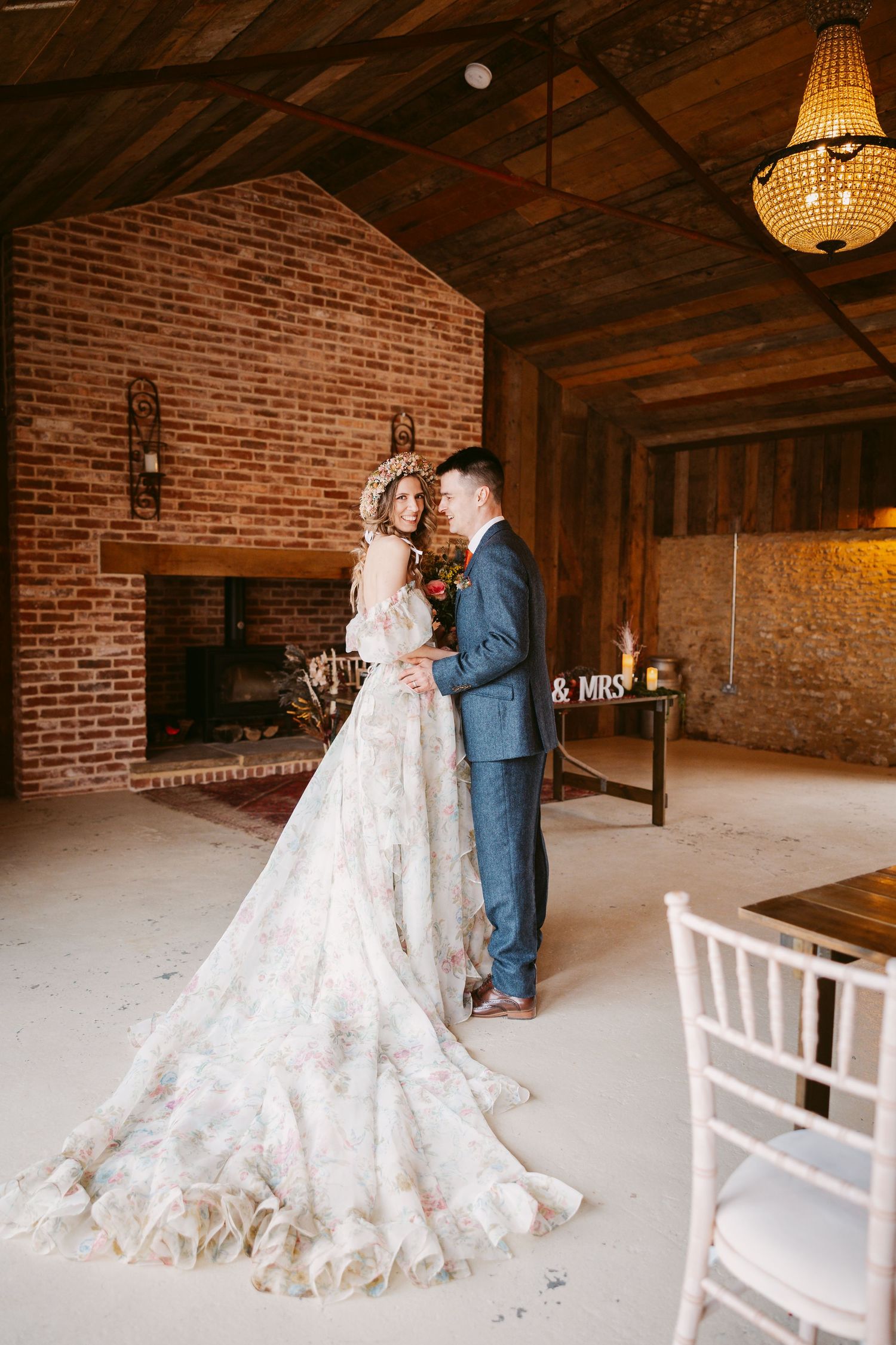 Romantic rustic wedding scene with bride in flowing lace gown and groom in blue suit inside brick-walled venue.