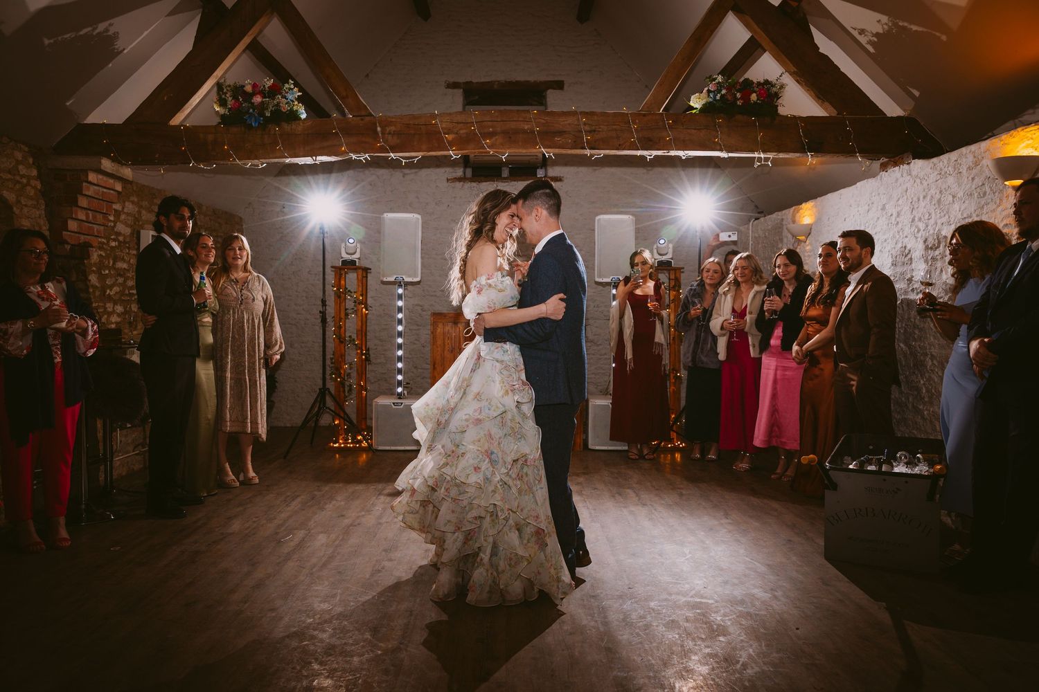 First dance at a wedding reception with guests watching in a rustic barn venue with exposed beams and warm lighting.