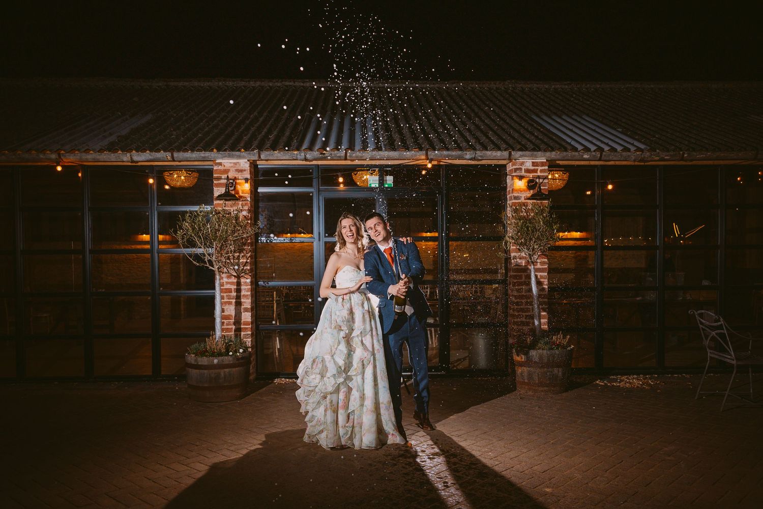 A couple shares a romantic moment outside a rustic venue with twinkling lights overhead.