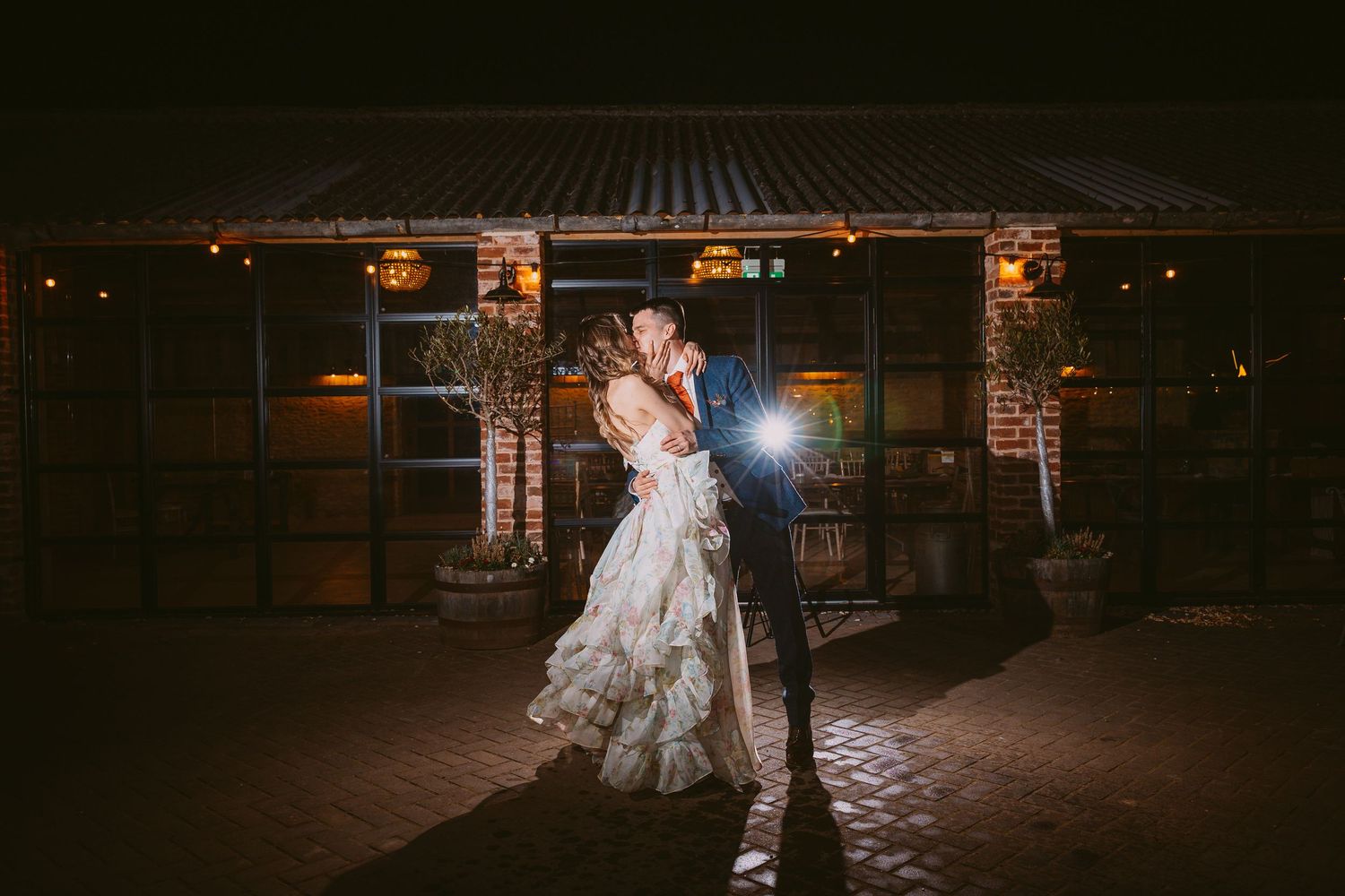 A couple embraces in an intimate dance pose outside a warmly lit rustic venue at night.