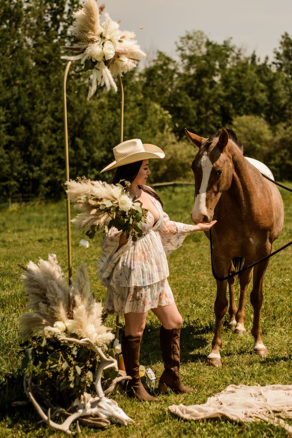 A brown horse stands in a grassy field with pampas grass and bohemian decorative elements.