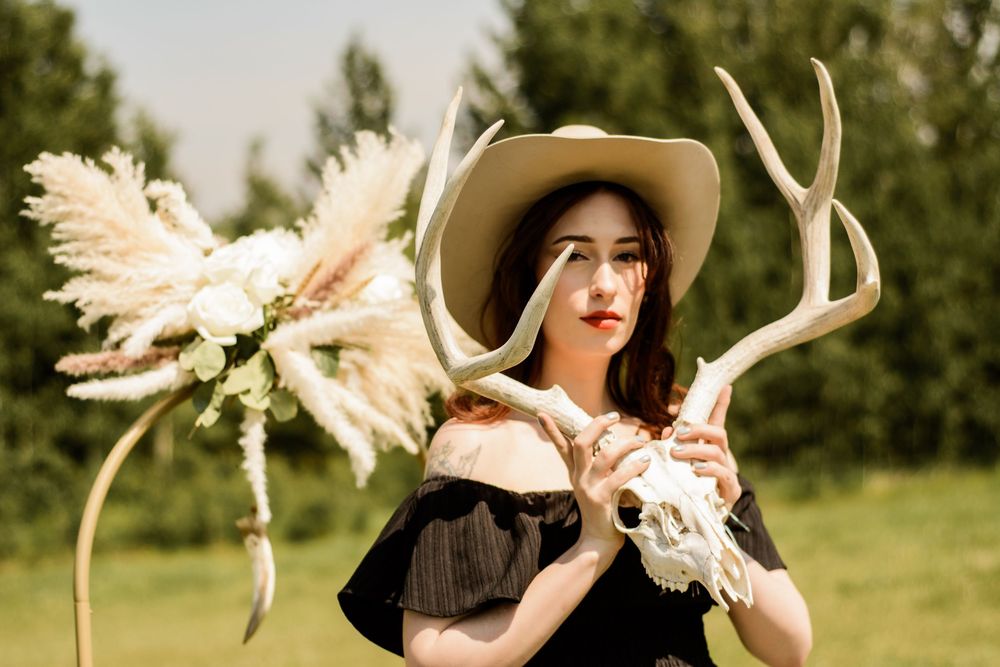 In an outdoor field setting, someone in a western hat holds a large deer skull with antlers and dried flowers.