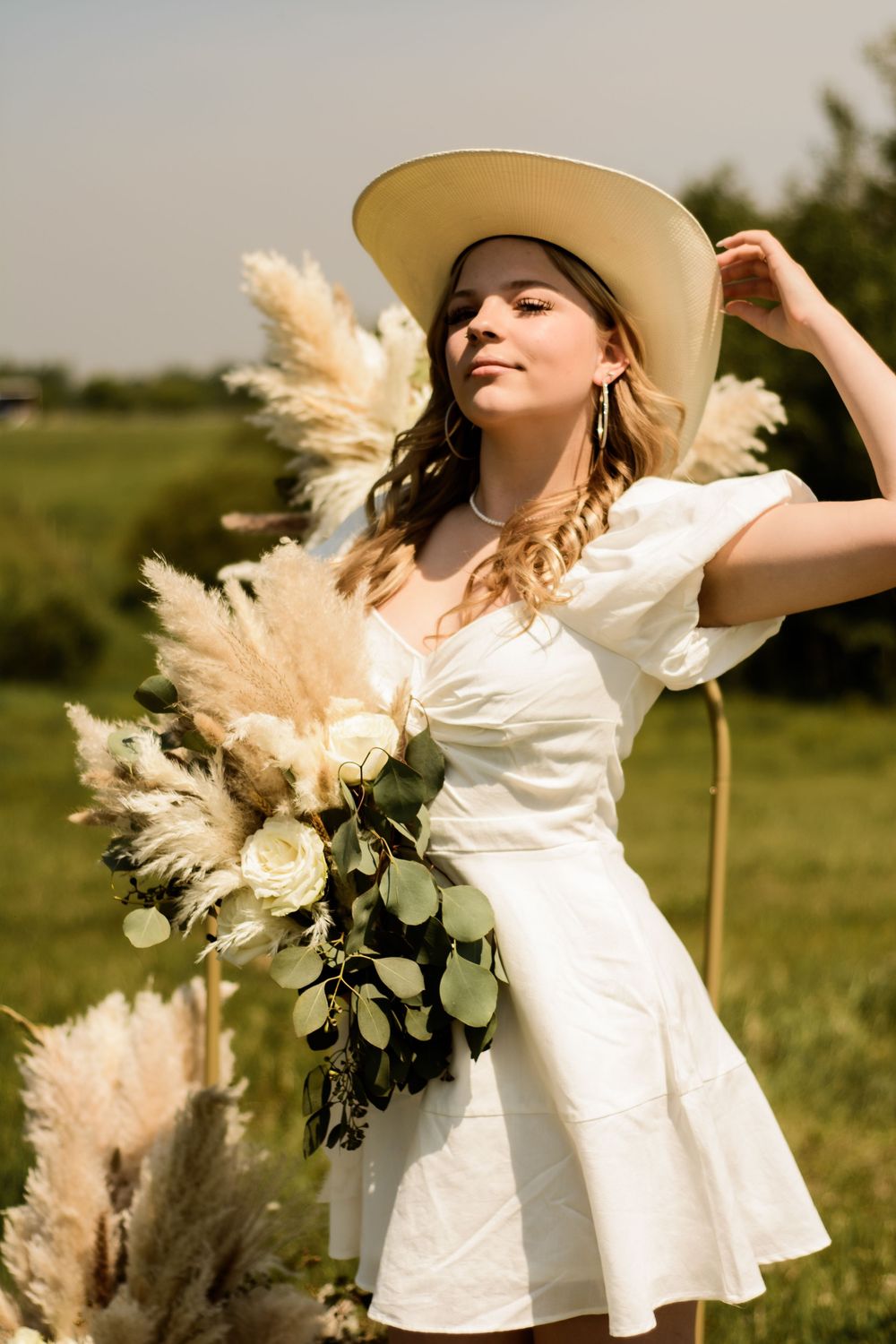 A dreamy outdoor portrait featuring a white sundress and wide-brimmed hat with pampas grass and rose bouquet.