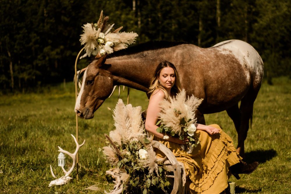 A person in a floral dress sits in a grassy field next to a brown and white horse among dried pampas grass arrangements.