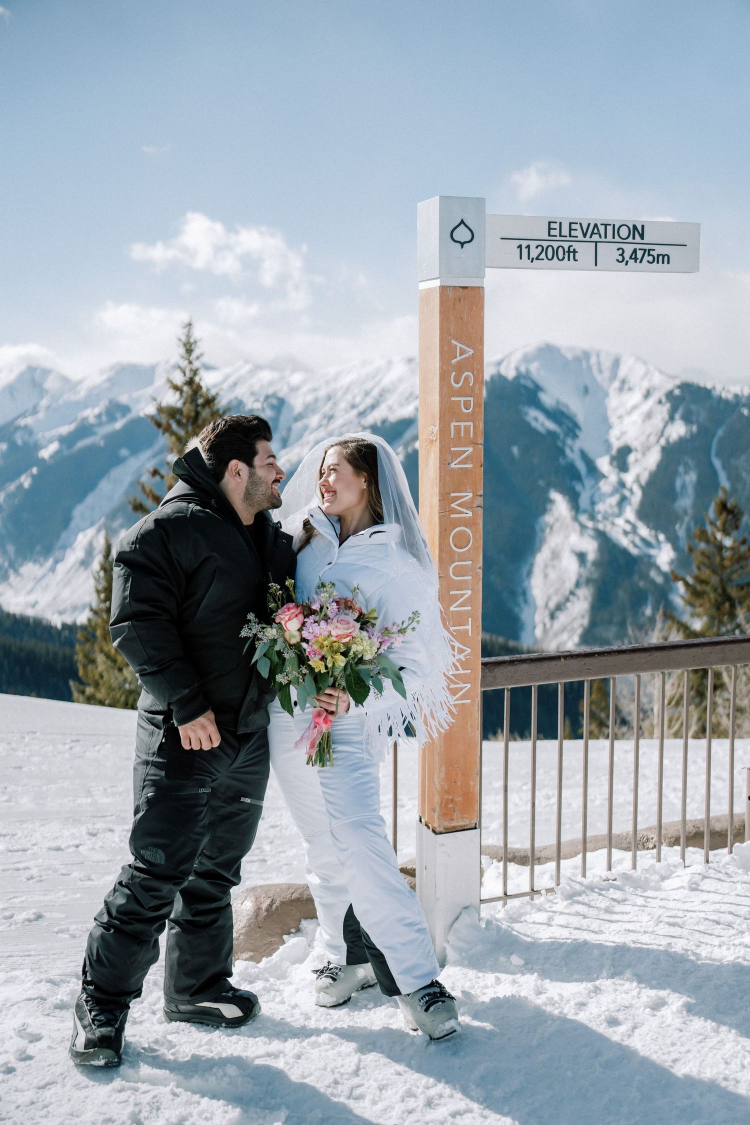Two people in winter ski gear share a moment by an elevation marker sign on a snowy mountain summit with alpine peaks behind.