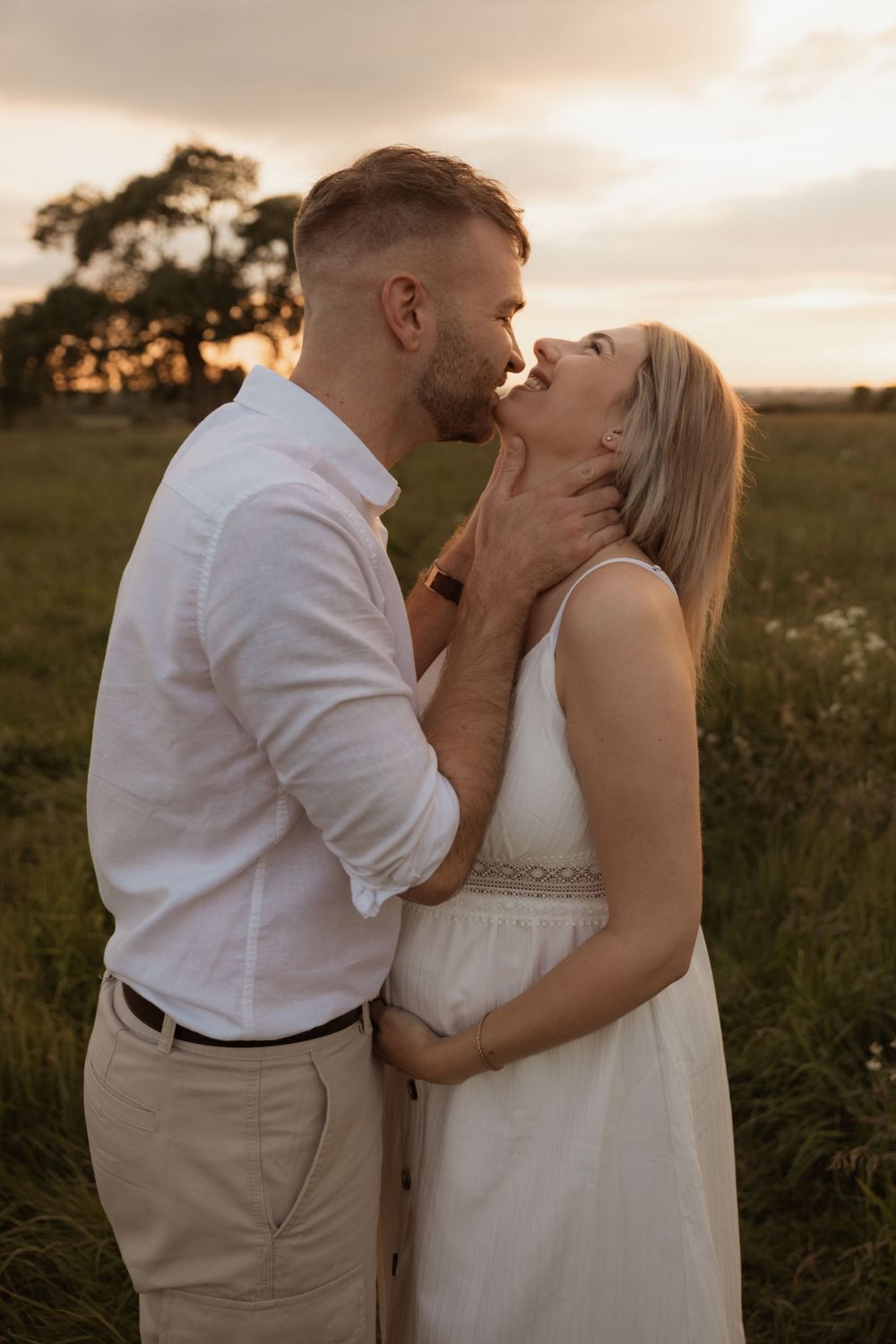 A romantic couple embraces in a grassy field at sunset, with the woman wearing a white dress and man in light clothing.
