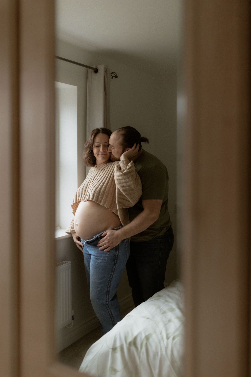 A tender moment captured through a doorway shows an expectant couple embracing near a window in a bedroom.