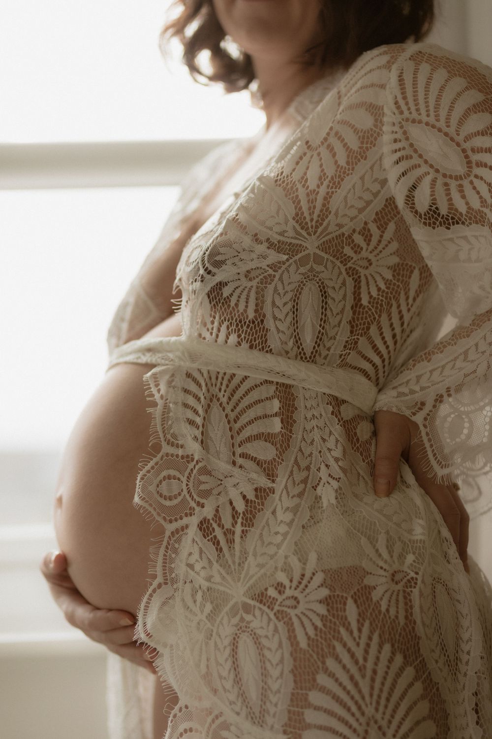 A pregnant belly draped in delicate white lace maternity dress with intricate floral patterns shows in soft window light.