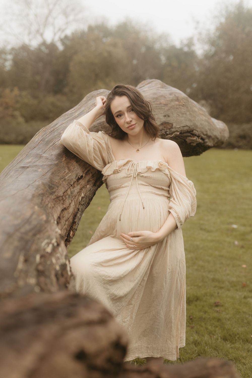 Expectant figure in a flowing cream dress poses beside a fallen tree trunk in a misty outdoor setting.