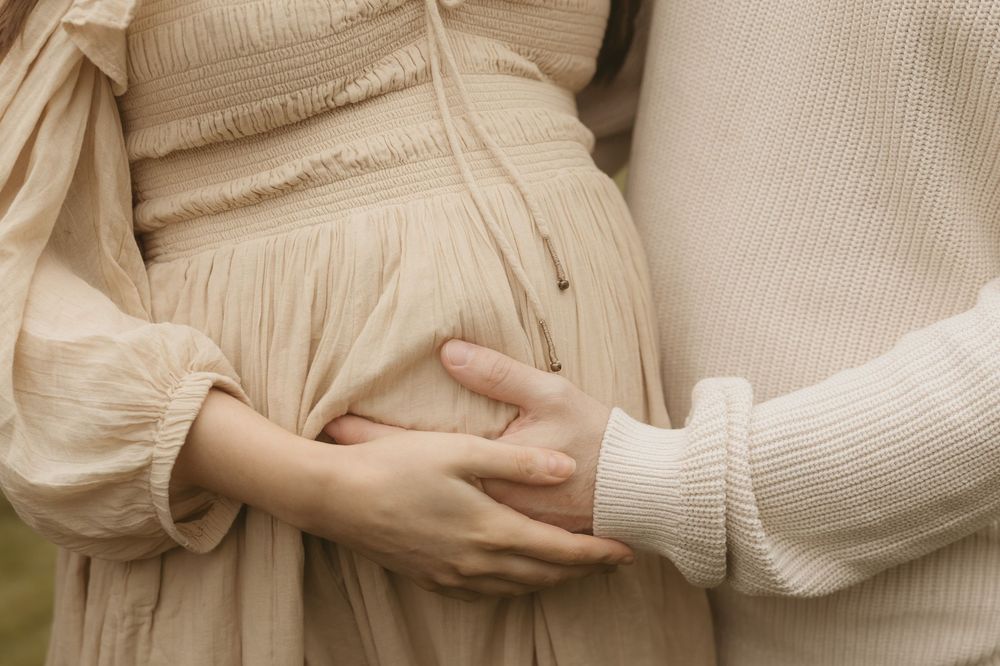Gentle hands touch a cream-colored maternity dress in soft natural light during a romantic pregnancy photoshoot.