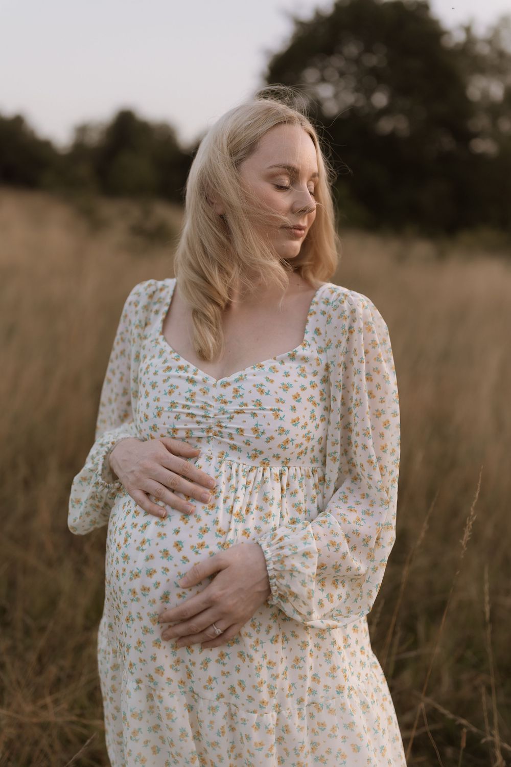 A pregnant woman in a floral dress stands in a golden field at sunset while gently touching her baby bump.