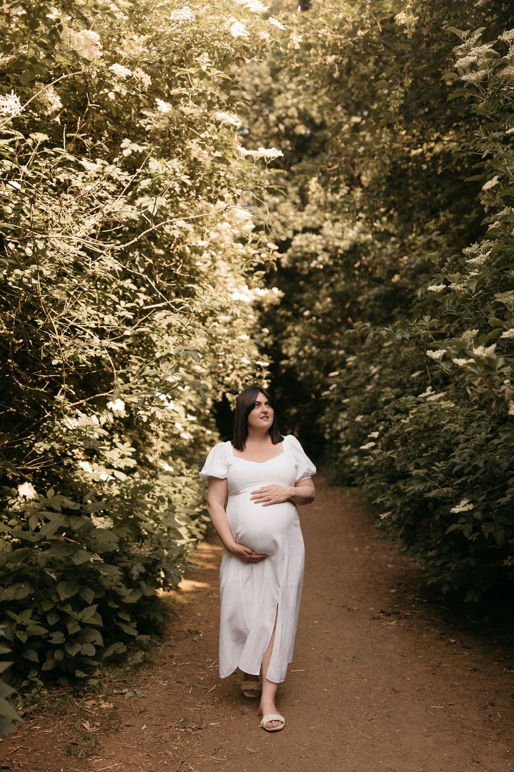 A person in a white summer dress sitting on a dirt path surrounded by lush greenery at golden hour.