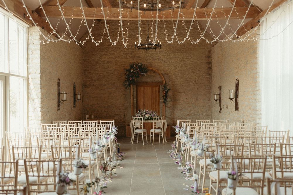 A rustic indoor wedding venue with white chairs, stone walls, string lights and floral arch at the altar.