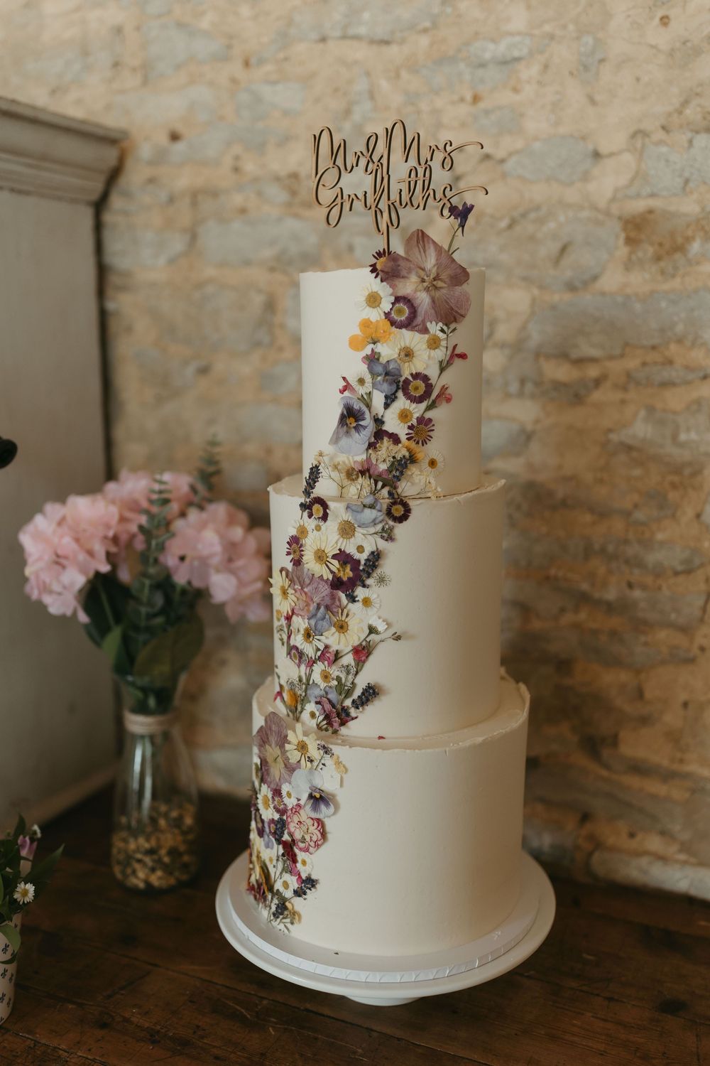 Elegant four-tier white wedding cake decorated with cascading dried flowers and modern calligraphy topper.